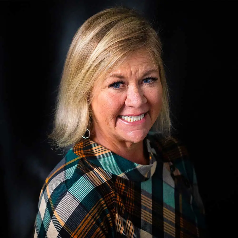 Blonde woman smiling at camera wearing teal and brown plaid shirt against dark background