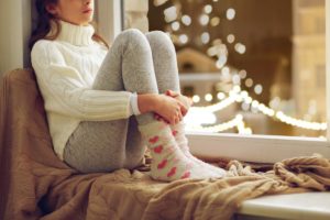 woman sitting forlornly in the window sill of large bay window dressed in white turtleneck and festive socks thinking about mental health and the holidays