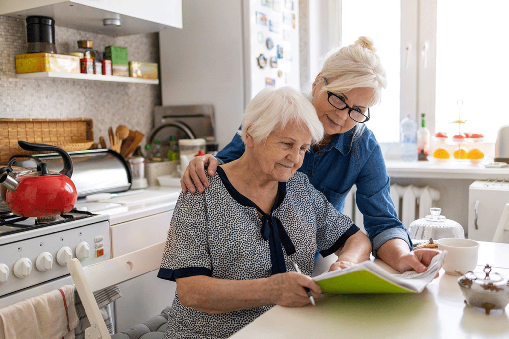 middle-aged woman standing over her elderly mother at kitchen table helping her with a memory game as part of National Alzheimer's Awareness Month
