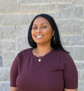 Woman with dark hair wearing burgundy top with pendant necklace smiling against stone wall