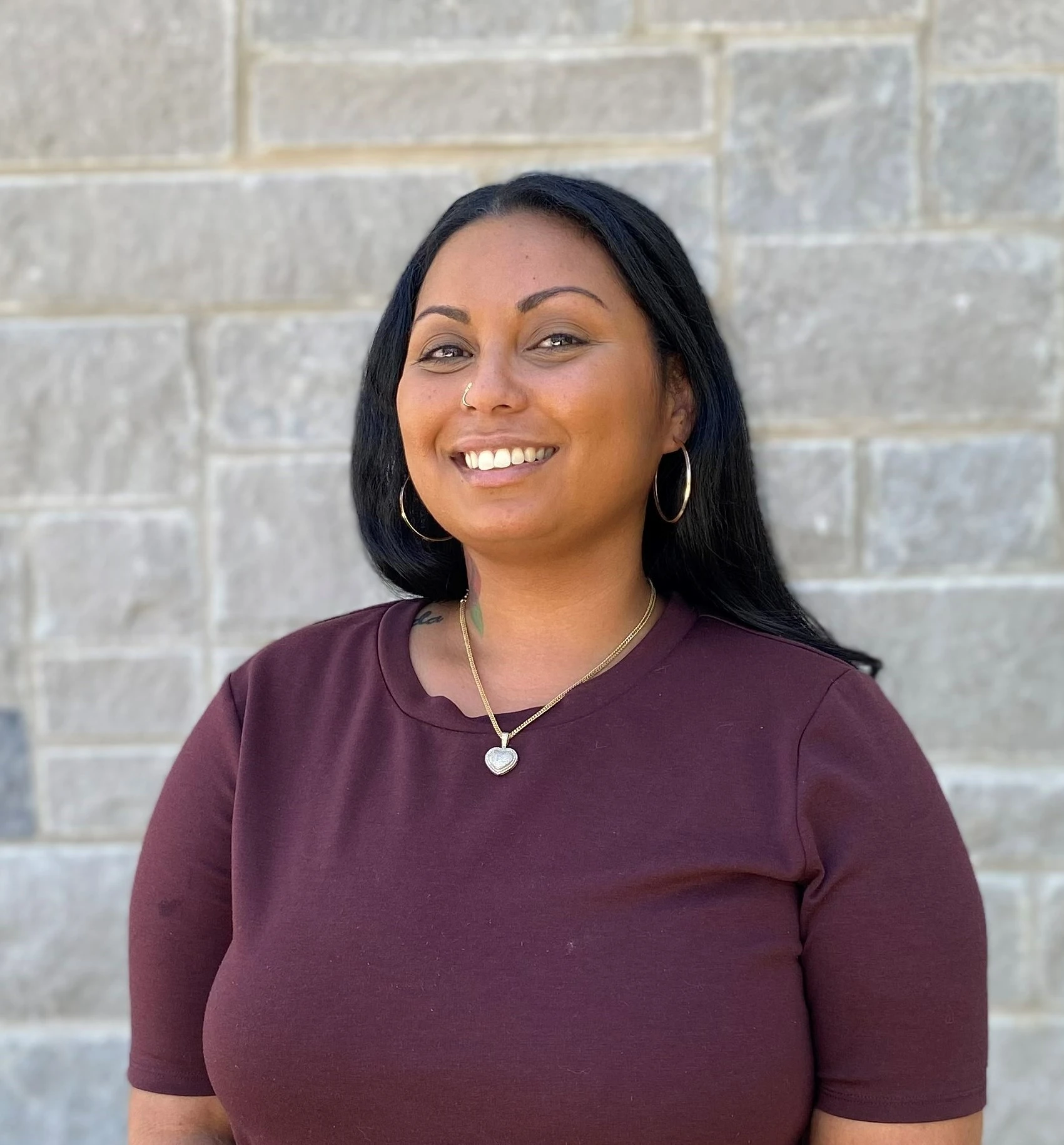 Woman with dark hair wearing burgundy top with pendant necklace smiling against stone wall