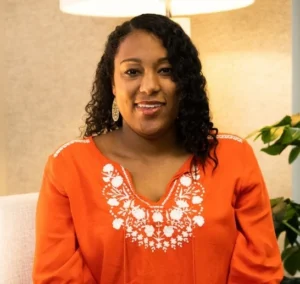 Woman with curly dark hair wearing bright orange top with white embroidered neckline, smiling indoors