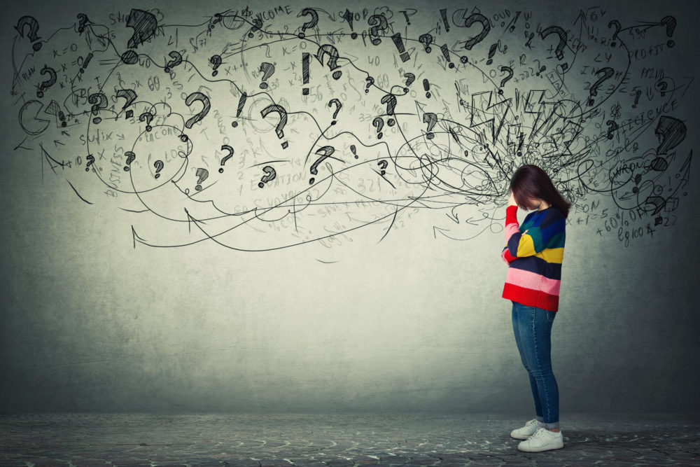 young woman standing in front of a mural of question marks with her head in her hand indicating she is worried and stressed with questions