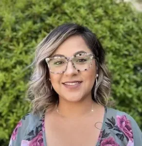 Woman with shoulder-length wavy blonde hair wearing clear-framed glasses and a floral patterned top, smiling at camera.