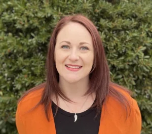 Woman with reddish-brown hair wearing orange cardigan over black top smiling at camera outdoors