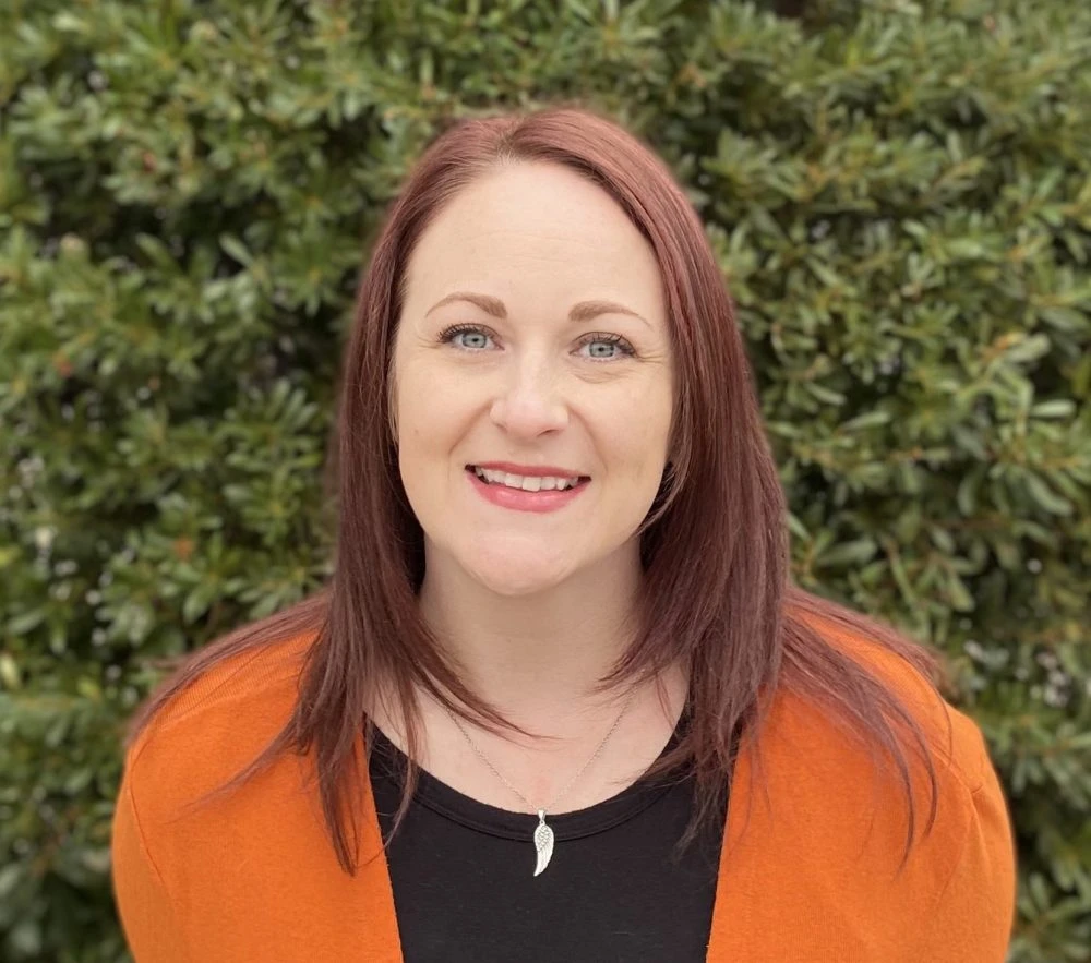Woman with reddish-brown hair wearing orange cardigan over black top smiling at camera outdoors