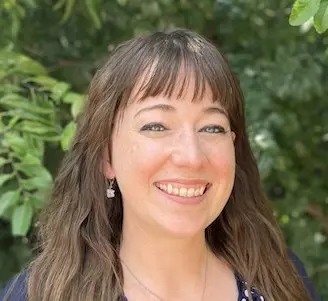 Woman with long brown wavy hair smiling at camera wearing dark top against green foliage background
