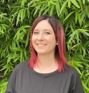 Woman with pink-tipped black hair wearing dark shirt smiling at camera with green foliage