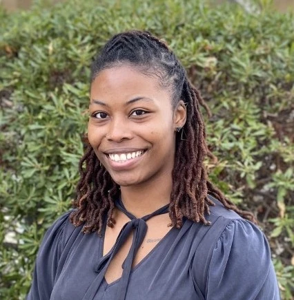 Woman with dark hair in braids wearing a dark gray button-up top with black necklace, smiling at camera against green foliage