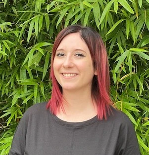 Woman with pink-tipped black hair wearing dark shirt smiling at camera with green foliage