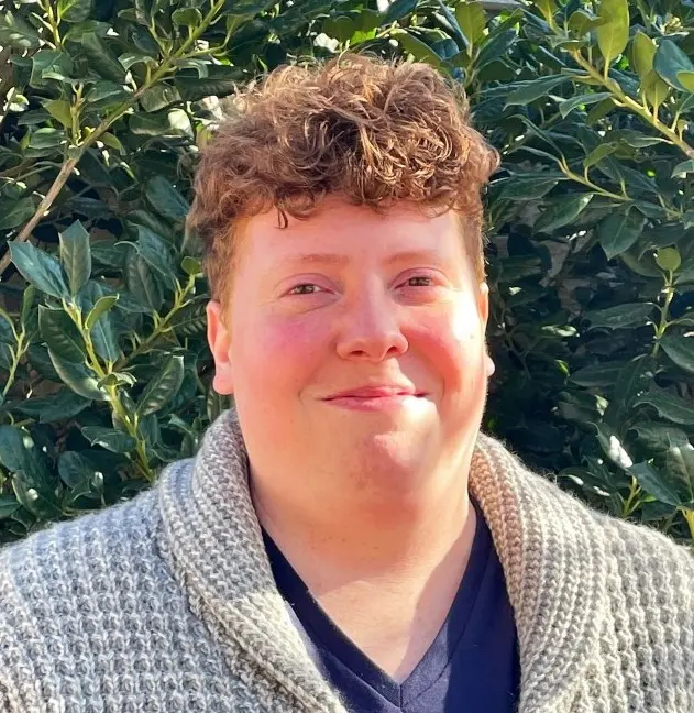 Person with reddish curly hair smiling at camera wearing navy v-neck shirt against green foliage background
