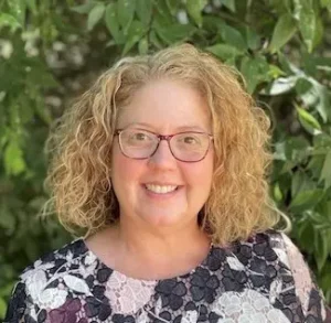 Woman with curly reddish-blonde hair wearing glasses and a patterned top with green foliage background