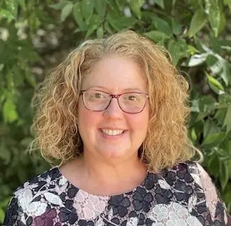 Woman with curly reddish-blonde hair wearing glasses and a patterned top with green foliage background