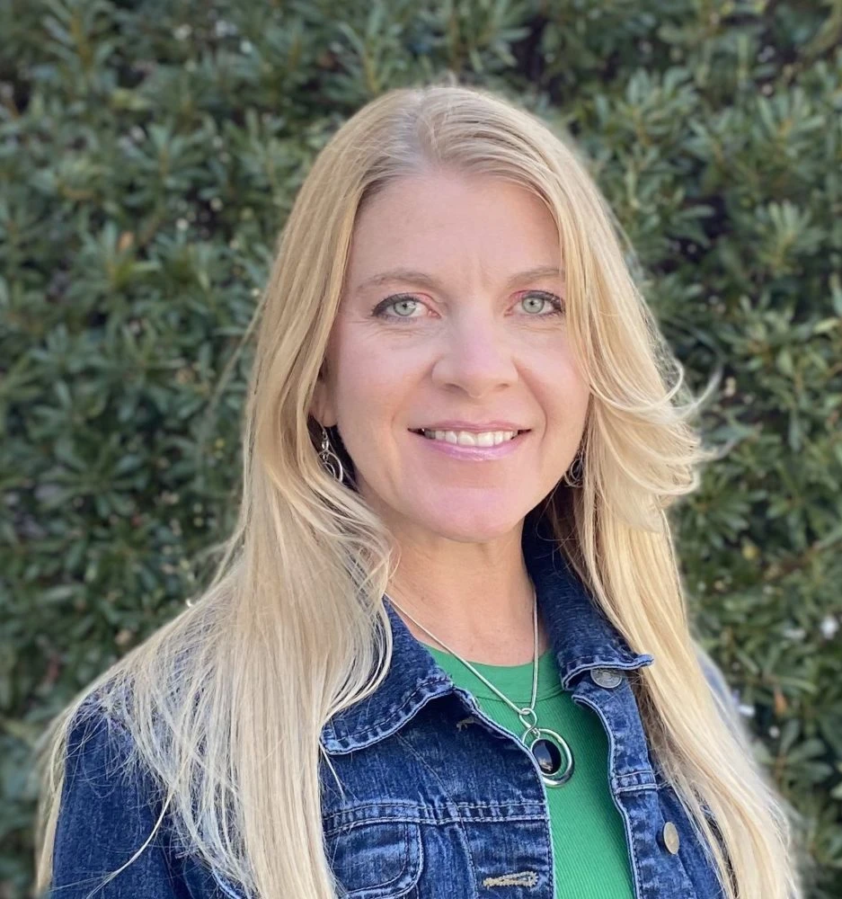 Woman with long blonde hair wearing a denim jacket over a green shirt, smiling at the camera with green foliage in the background.