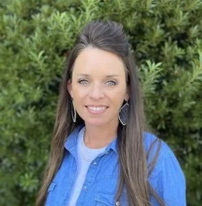Woman with long dark hair styled in an updo wearing a blue jacket and silver hoop earrings, smiling outdoors