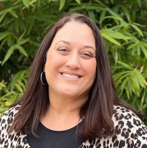 Woman with long dark hair wearing hoop earrings and a black top with a leopard print pattern, smiling at the camera outdoors.