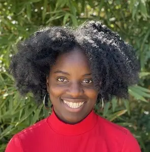 Woman with dark curly hair wearing a red top, smiling outdoors with green foliage in soft focus behind her