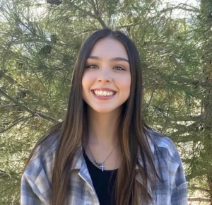 Woman with long dark hair and necklace wearing plaid shirt smiling at camera with trees
