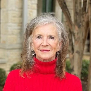 An older woman with long gray and white hair smiling gently at the camera, wearing a bright red turtleneck sweater, photographed against a blurred ...