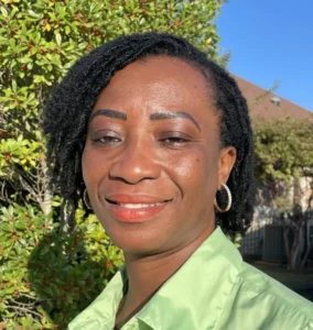 A woman with short dark curly hair wearing gold hoop earrings and a lime green shirt outdoors with mountains