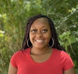 Woman with dark braided hair wearing red t-shirt, smiling at camera with green foliage background