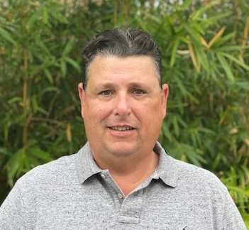 Man with dark hair smiling at camera, wearing gray polo shirt against green foliage background