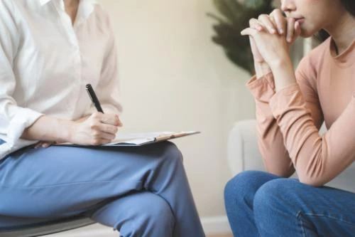 Therapist taking notes while listening to a patient in blue jeans during a counseling session indoors