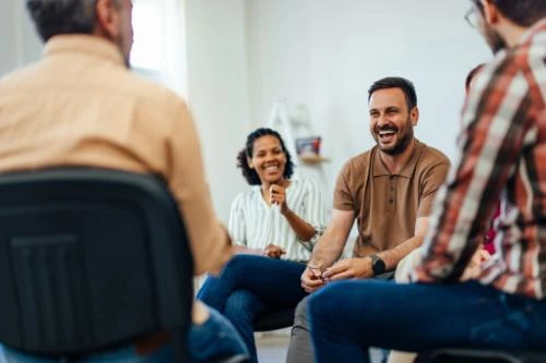 Group of diverse people sitting in a circle during what appears to be a support meeting or group discussion indoors.