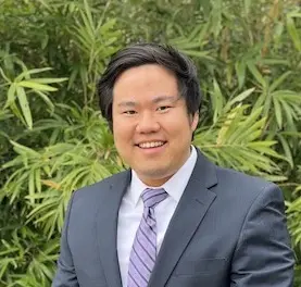 Professional headshot of an Asian man in a gray blazer and purple striped tie, smiling at the camera with green foliage in the background