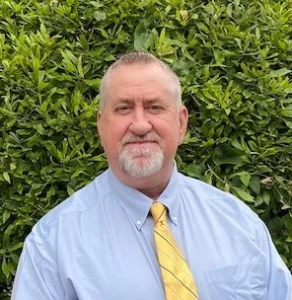 Older man with gray hair and beard wearing a light blue shirt and yellow tie, smiling outdoors against green foliage background.