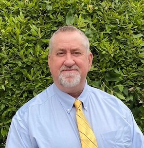Older man with gray hair and beard wearing a light blue shirt and yellow tie, smiling outdoors against green foliage background.