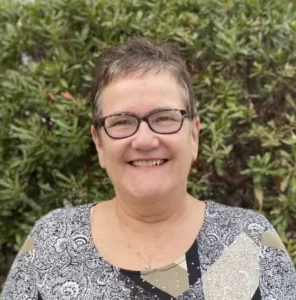 Woman with short dark hair wearing glasses and gray top, smiling outdoors with green foliage in background
