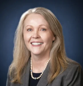 A professional headshot of a woman with long light brown hair wearing a gray blazer and pearl necklace, smiling against a dark background