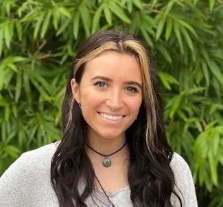 Young woman with long dark hair wearing a white sweater and a pendant necklace, smiling outdoors with green foliage behind her.