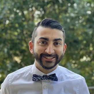 A portrait of a man with dark hair and beard wearing a white dress shirt with a patterned bow tie, smiling outdoors