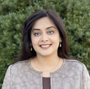 Woman with long dark hair and earrings wearing a gray top with dark center panel smiling with green background