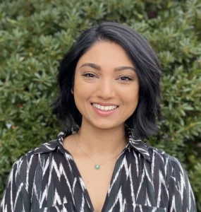 Smiling woman with shoulder-length dark hair wearing patterned dark and white shirt, with green foliage background