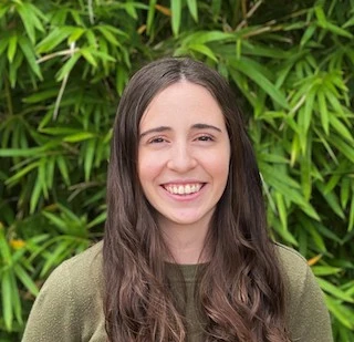 Young woman with long dark hair smiling at camera against green foliage background, wearing olive green shirt