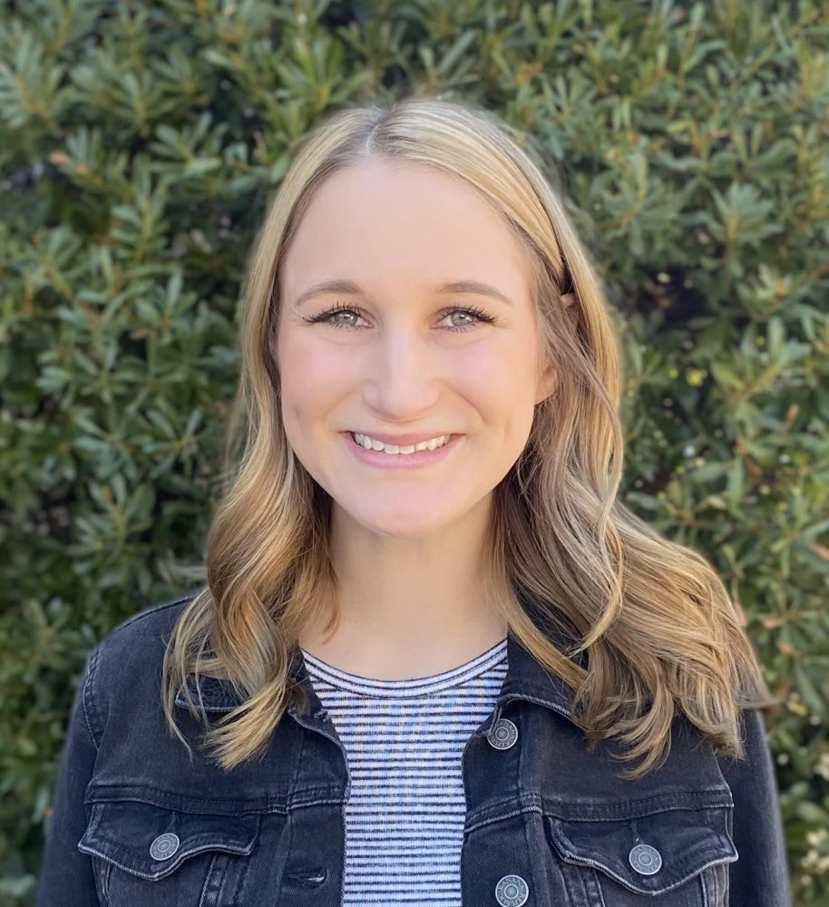 Smiling woman with long wavy brown hair wearing a denim jacket over a striped shirt against a green hedge