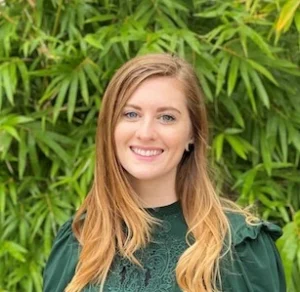 Young woman with reddish-blonde hair smiling at camera wearing dark green shirt against green foliage background