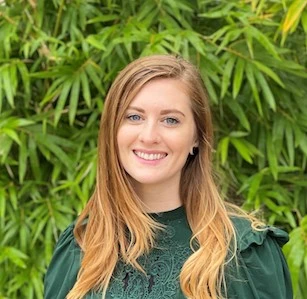 Young woman with reddish-blonde hair smiling at camera wearing dark green shirt against green foliage background