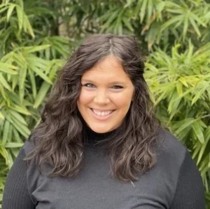 Woman with long dark hair wearing a black ribbed top, smiling at camera with green foliage behind