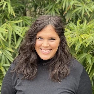 Woman with long dark hair wearing a black ribbed top, smiling at camera with green foliage behind