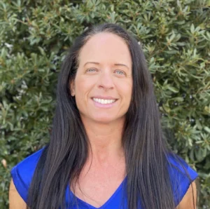 Woman with long dark hair wearing bright blue top, smiling at camera against green foliage background
