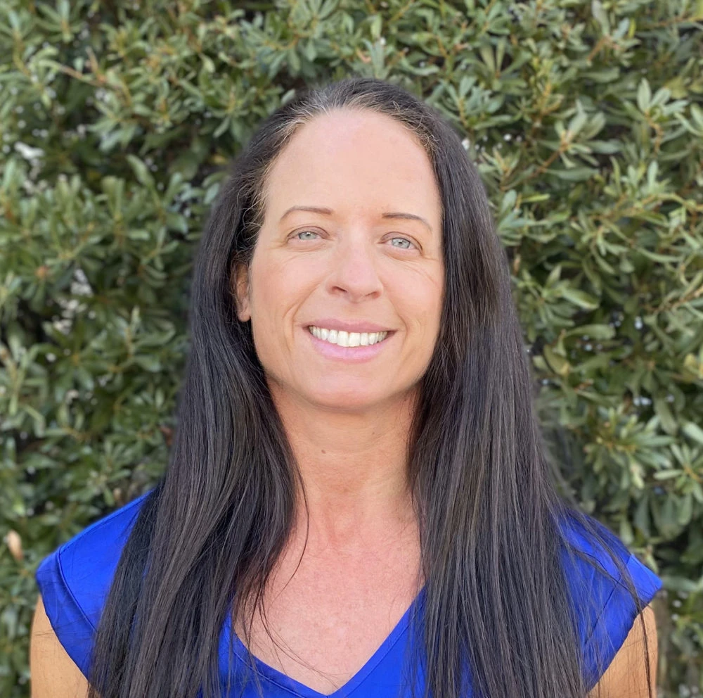 Woman with long dark hair wearing bright blue top, smiling at camera against green foliage background