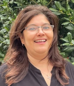 A woman with long reddish-brown hair wearing glasses and a black top smiling outdoors with green foliage