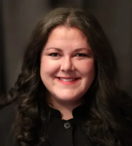 Professional headshot of a woman with long dark wavy hair and red lipstick, wearing black clothing, photographed against a dark blurred background.
