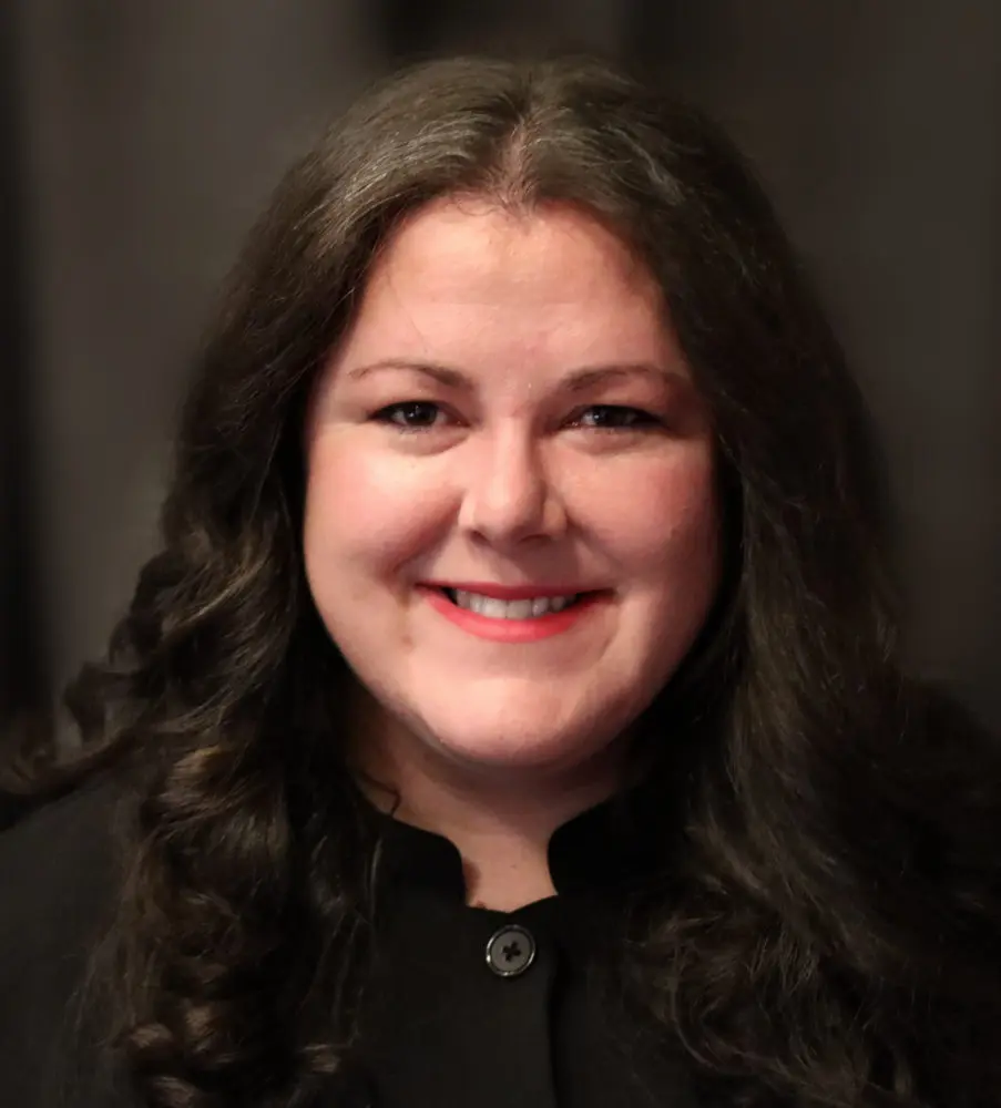 Professional headshot of a woman with long dark wavy hair and red lipstick, wearing black clothing, photographed against a dark blurred background.