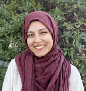 Woman wearing a burgundy hijab and white top, smiling at camera against a blurred green background.