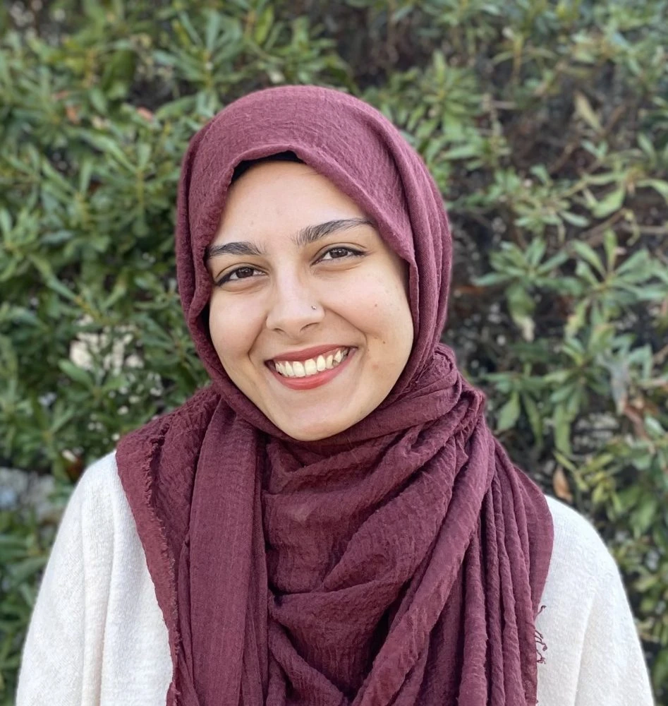 Woman wearing a burgundy hijab and white top, smiling at camera against a blurred green background.